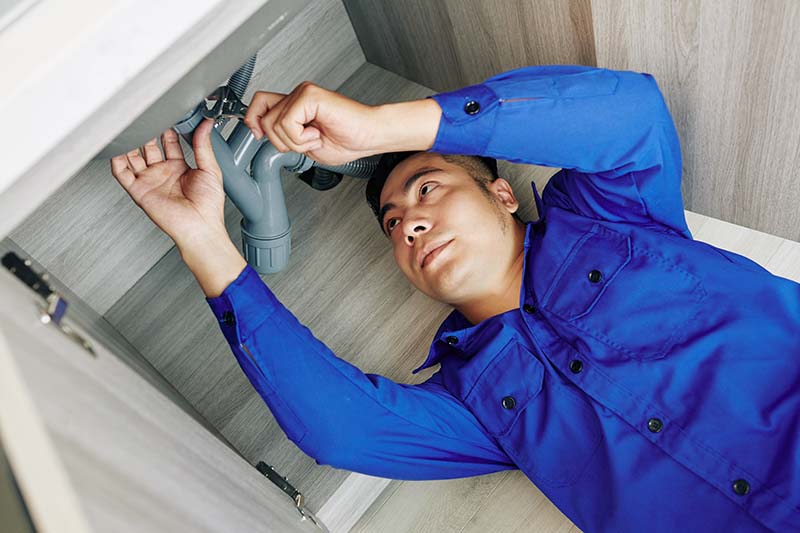 A plumber working under a sink wearing blue overalls