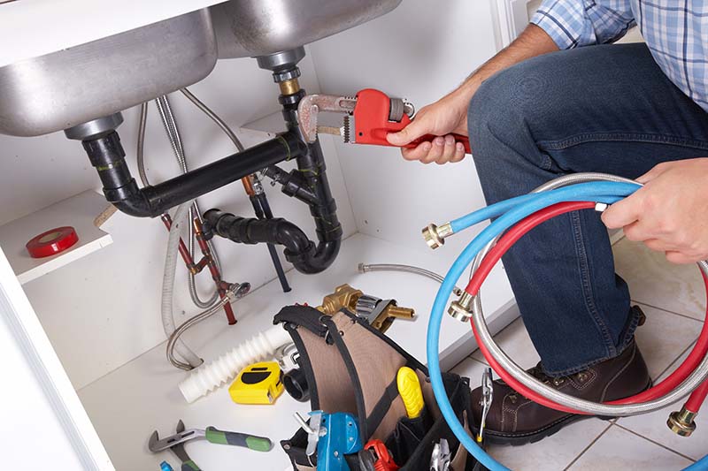 A person holding a wench and washing machine pipes in front of a kitchen sink cupboard